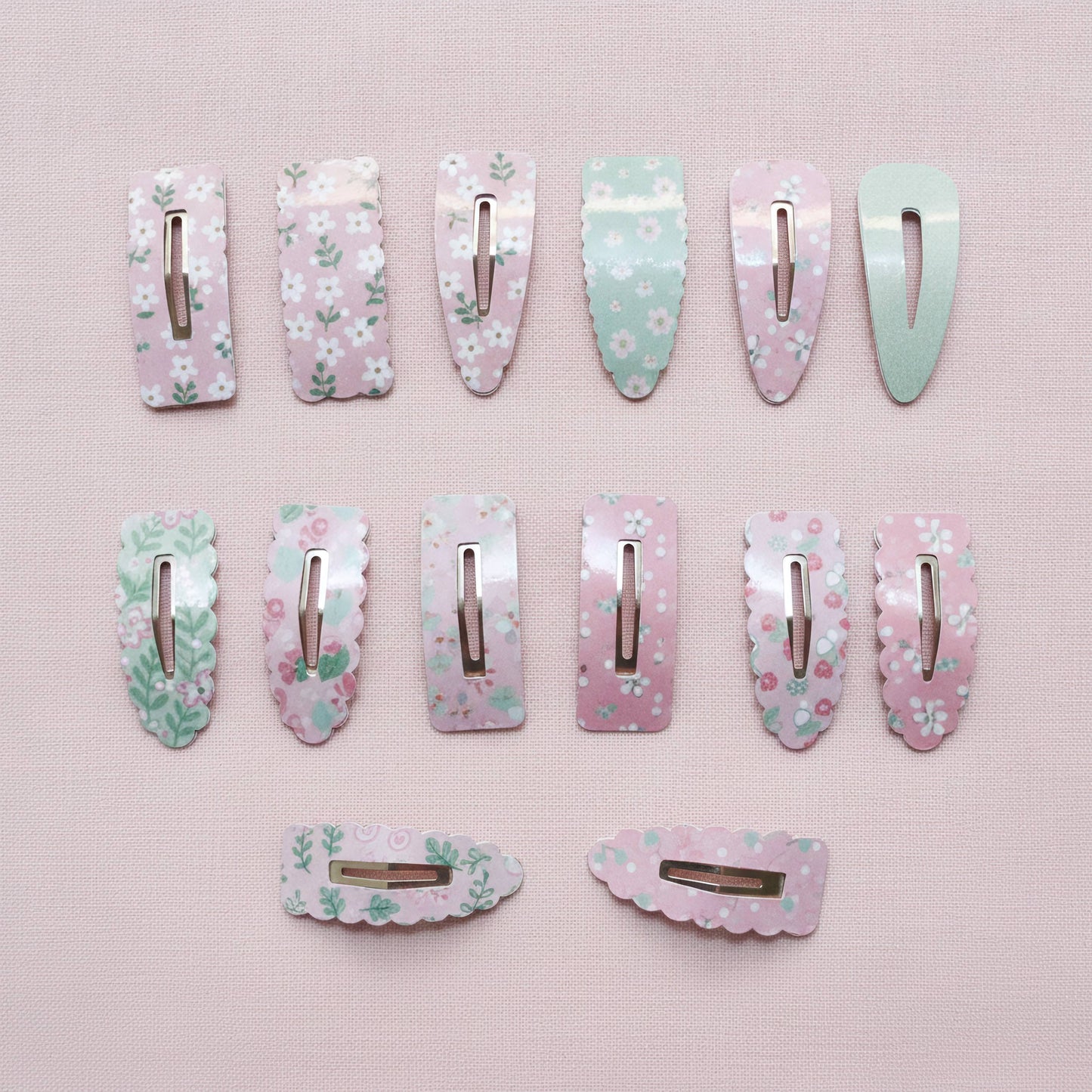 Twelve pastel floral hair clips arranged on a light pink textured background, including several long rectangular clips and two shorter curved clips. The clips feature pink, mint green, and white floral patterns with shiny metallic silver clasps.