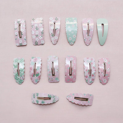 Twelve pastel floral hair clips arranged on a light pink textured background, including several long rectangular clips and two shorter curved clips. The clips feature pink, mint green, and white floral patterns with shiny metallic silver clasps.
