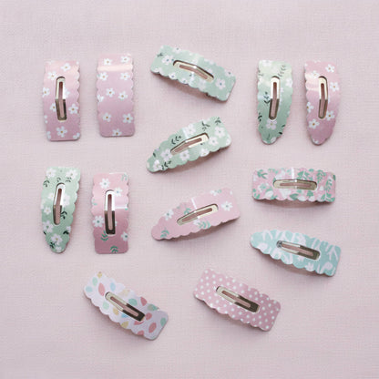A set of twelve pastel hair claw clips with various floral and polka dot patterns in pink, mint green, and light blue, arranged on a soft pink background.