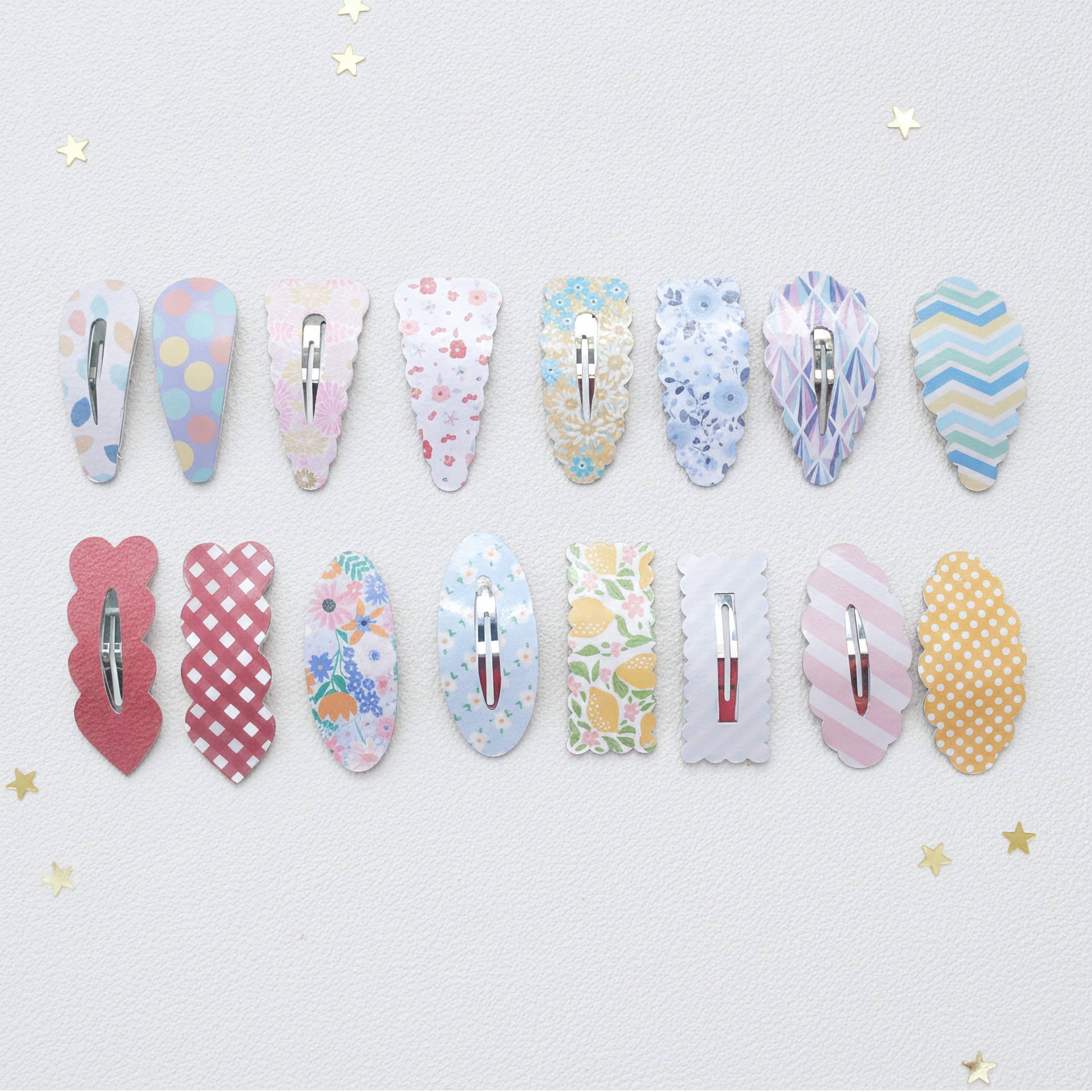 Twelve decorative hair claw clips arranged in two rows on a light textured background with small gold star accents. The clips feature various pastel patterns including polka dots, florals, stripes, and chevron designs in pink, blue, yellow, and white.