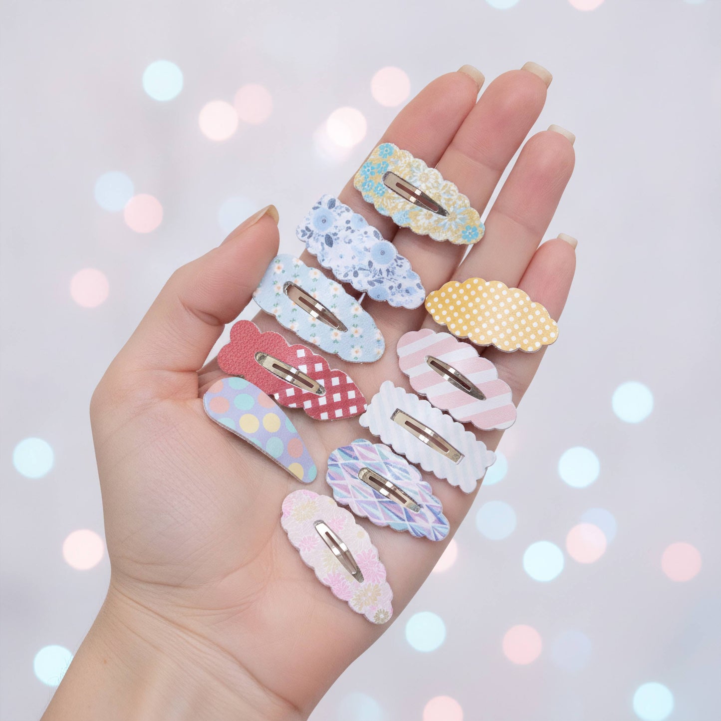 A hand holds a collection of nine pastel-colored hair clips with various patterns, including polka dots, stripes, and florals, set against a soft-focus background with colorful bokeh lights.