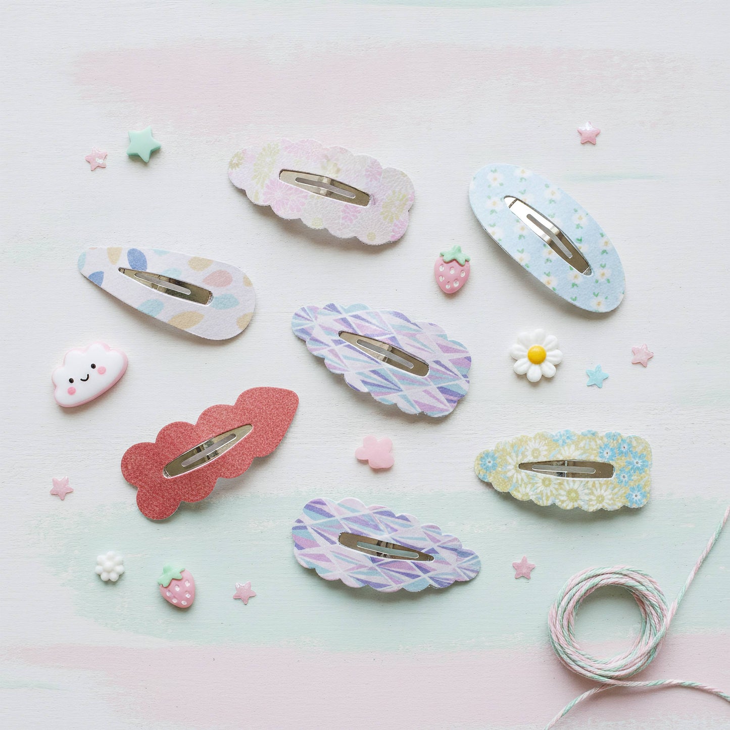 Six pastel hair claw clips arranged on a light pastel pink and blue background with small decorative paper stars, flowers, strawberries, and twine. The clips have assorted patterns including floral, striped, and polka dots in pink, blue, purple, and white.
