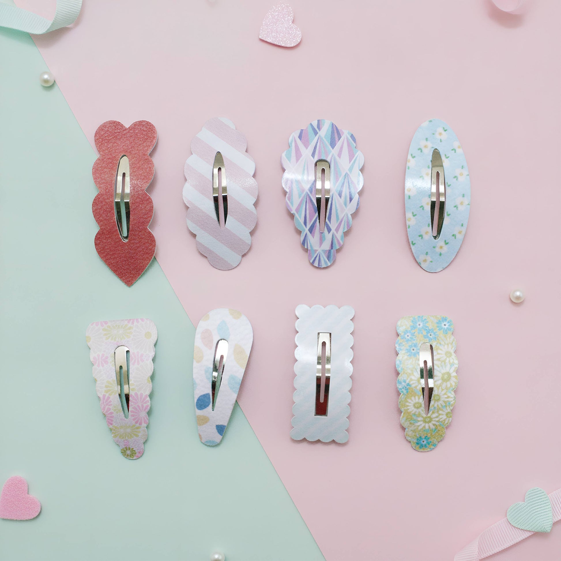 Eight hair claw clips in various pastel patterns and colors are arranged on a diagonally split pink and mint green background with small decorative hearts and pearls.