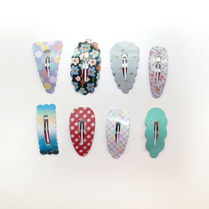 Eight decorative hair clips arranged in two rows on a plain white background. The clips have assorted pastel and bright patterns including polka dots, flowers, gingham, and solid colors, with silver metal spring clips visible at the back.