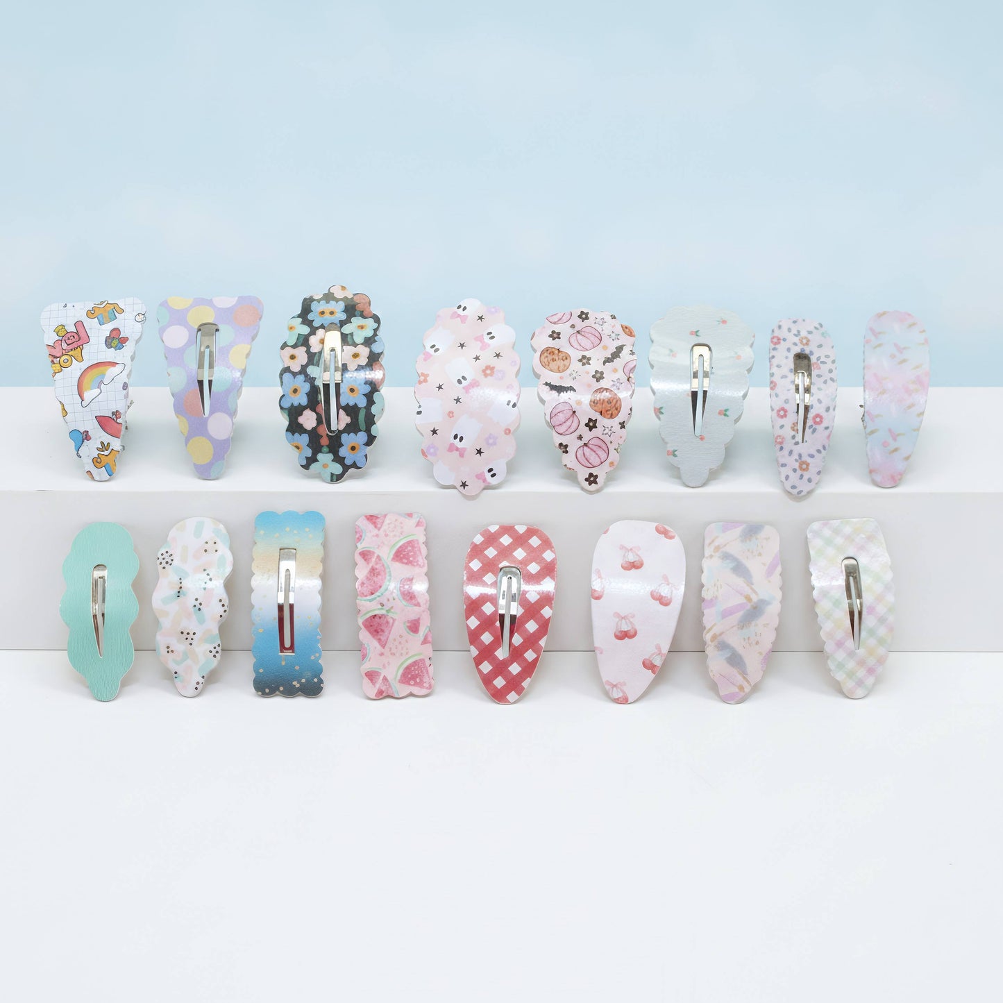 Twelve decorative hair clips arranged in two rows on a white display block against a pale blue background. The clips feature various pastel patterns including polka dots, florals, hearts, checkers, and watermelon prints.