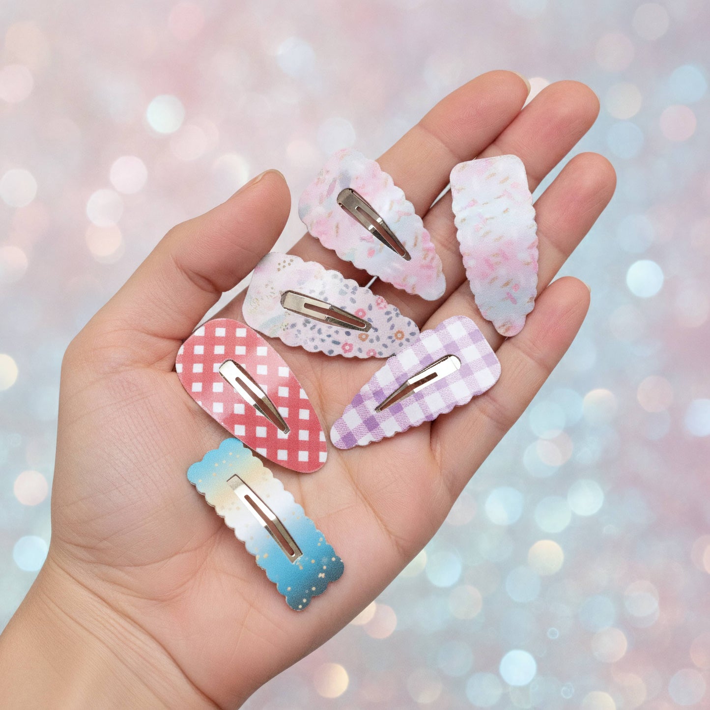 A hand holding six decorative hair claw clips in pastel colors and patterns, including pink floral, white lace, red gingham, lavender check, and blue gradient designs, against a soft pink glittery bokeh background.