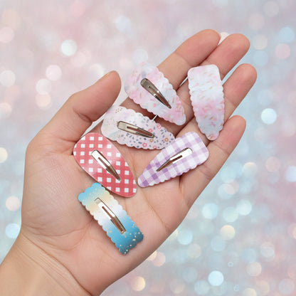 A hand holding six decorative hair claw clips in pastel colors and patterns, including pink floral, white lace, red gingham, lavender check, and blue gradient designs, against a soft pink glittery bokeh background.