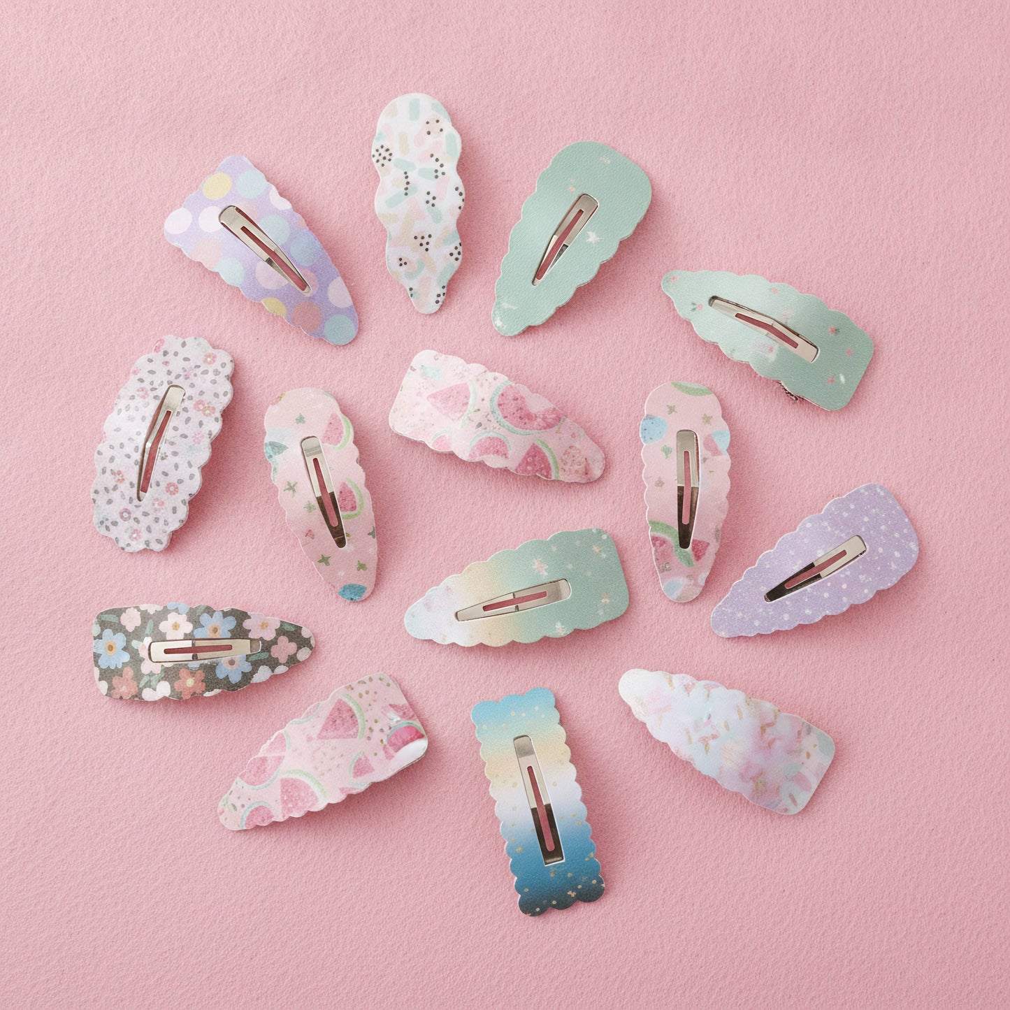 A set of pastel decorative hair clips arranged on a soft pink background. The clips feature various patterns including floral prints, polka dots, stars, and watercolor gradients in light blue, mint green, lavender, and pink, with rose gold metal clasps visible.