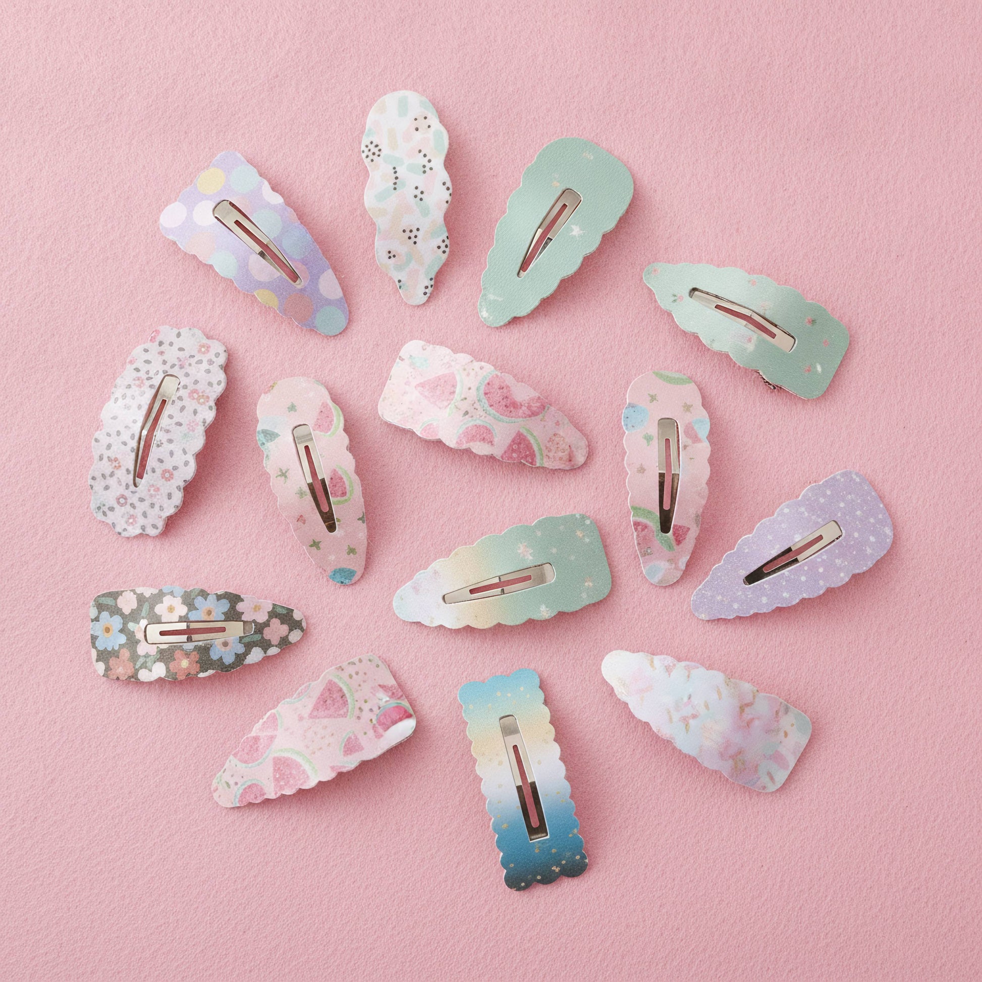 A set of pastel decorative hair clips arranged on a soft pink background. The clips feature various patterns including floral prints, polka dots, stars, and watercolor gradients in light blue, mint green, lavender, and pink, with rose gold metal clasps visible.