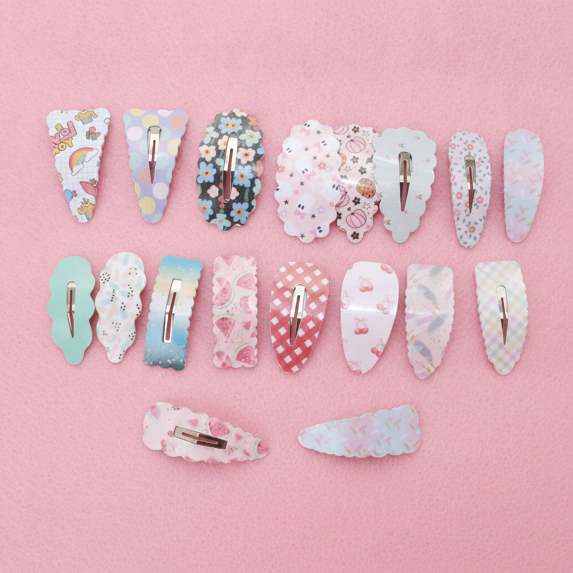 Twelve decorative hair claw clips arranged in rows on a solid pink background. The clips feature various pastel patterns including florals, checkers, stripes, and cute illustrations, with silver metal spring clips visible at the back.