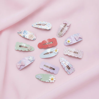 A set of ten pastel hair clips arranged on a soft pink fabric background. The clips feature various designs including floral prints, polka dots, glittery textures, bows, flowers, and cloud shapes in pastel colors like pink, blue, green, and purple.