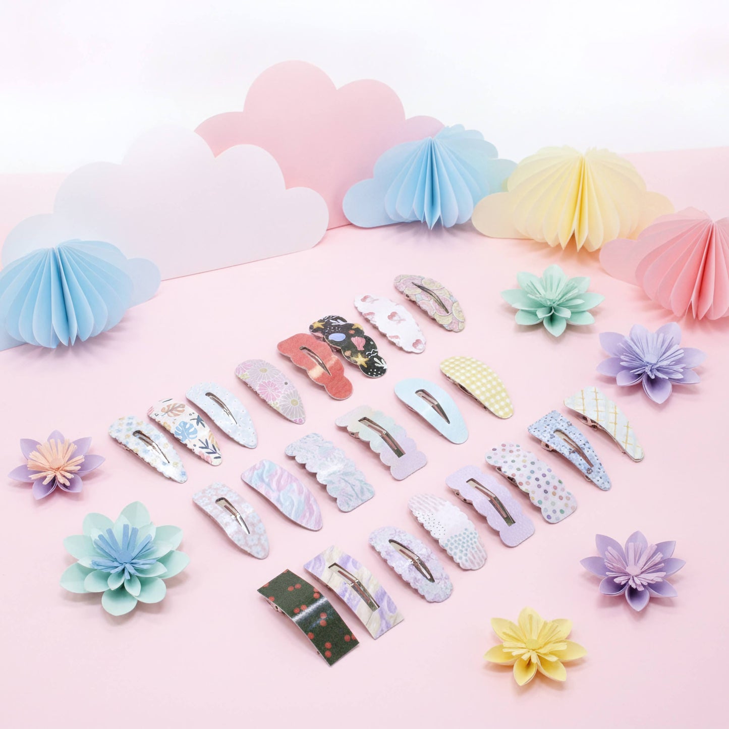 A collection of pastel hair claw clips arranged on a pink surface with decorative paper clouds and flowers in the background. The clips feature various patterns and colors including floral, polka dot, marble, and solid pastel designs.