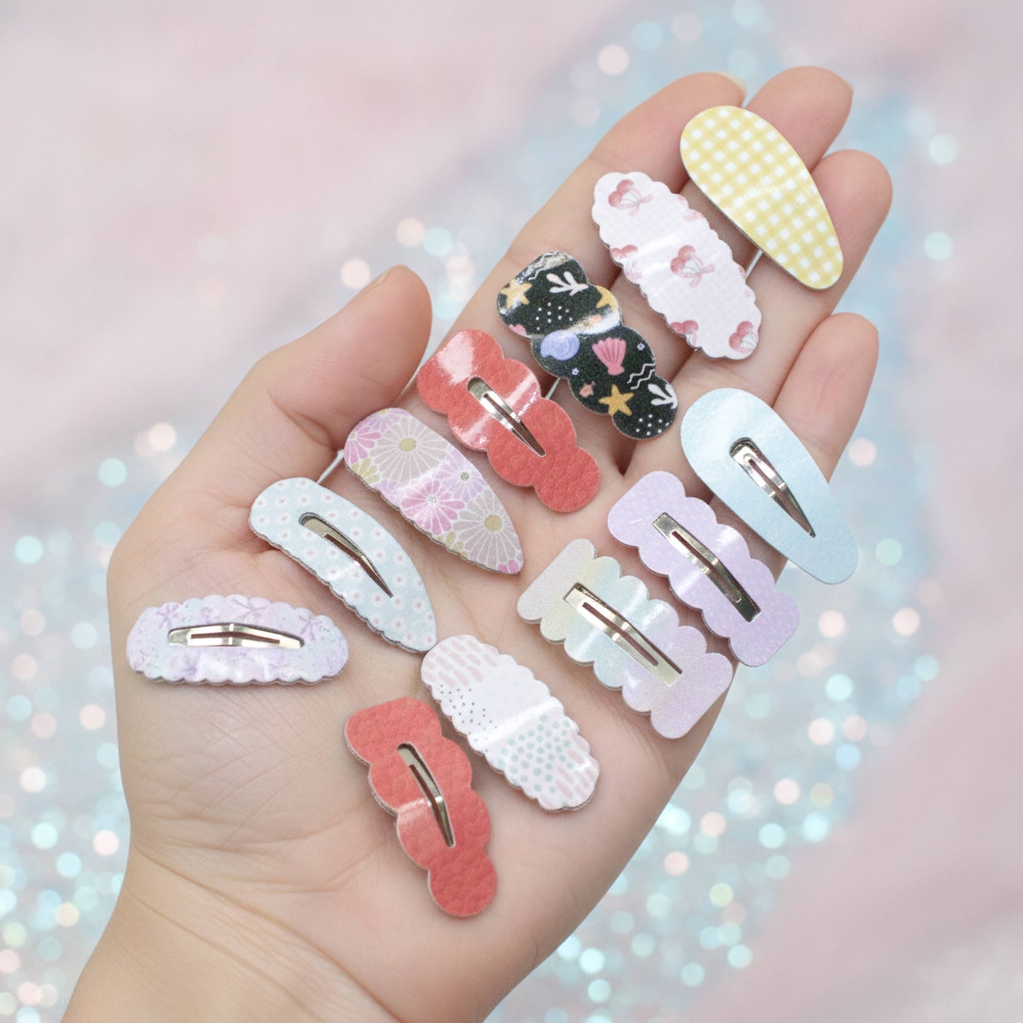 A hand holds multiple pastel hair claw clips in various patterns and colors, including pink, yellow, black, and white, against a soft pink bokeh background.