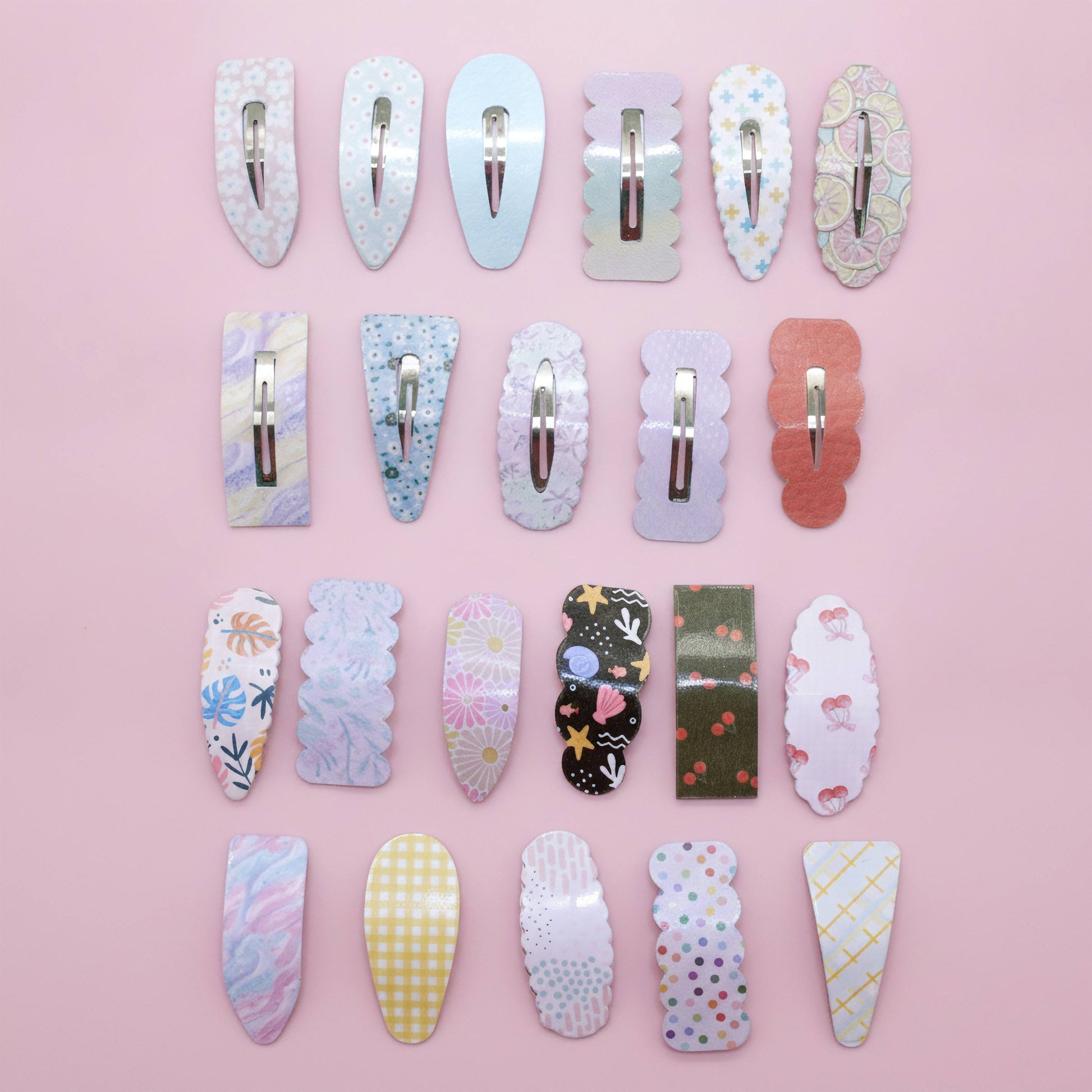 A set of 20 decorative hair clips arranged in a neat 4x5 grid on a solid pink background. The clips feature various pastel patterns including florals, stripes, polka dots, and gingham, with silver metal spring clips.