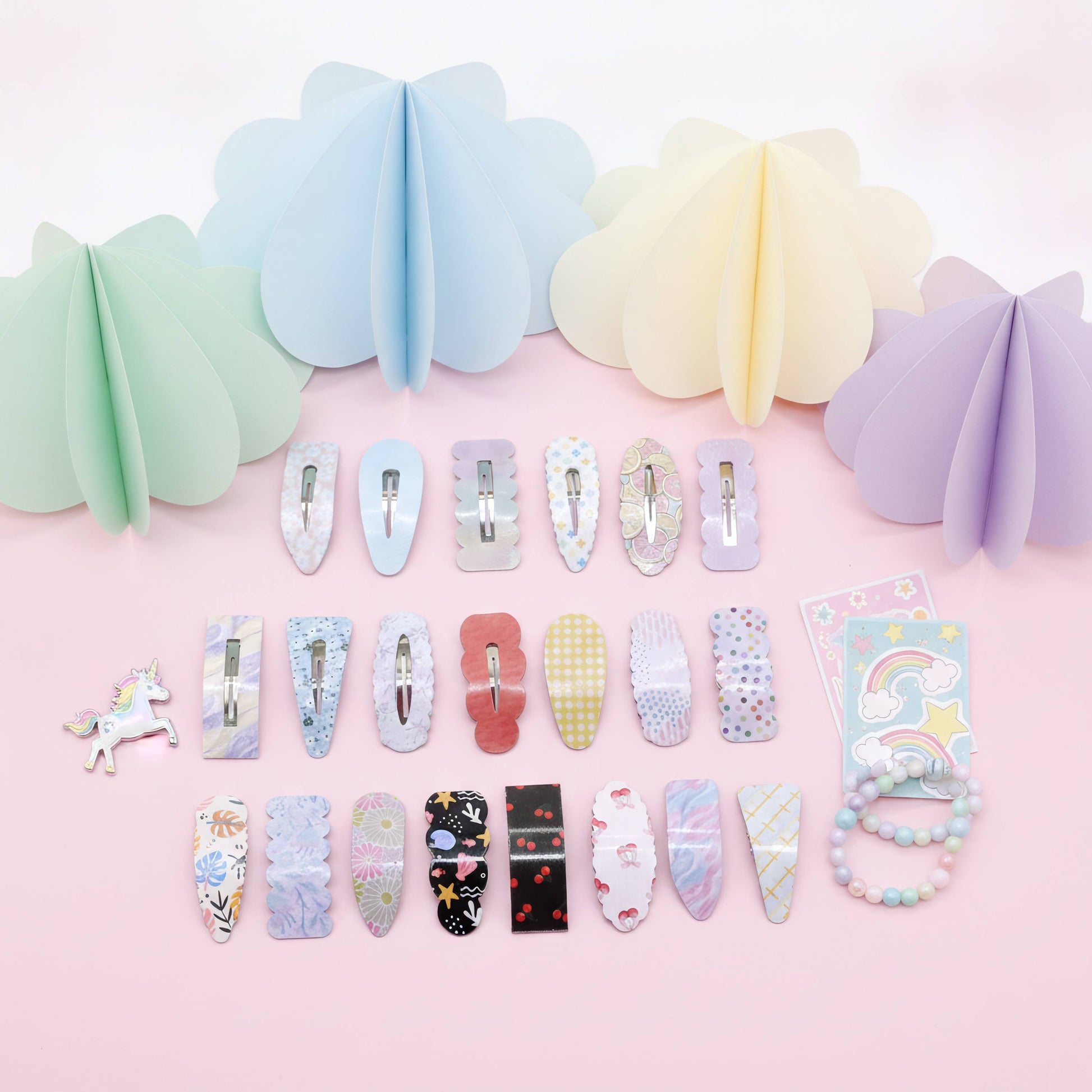 A collection of pastel hair claw clips and decorative hair accessories arranged on a light pink background, with matching paper fan decorations above. The clips feature various patterns including florals, stripes, polka dots, and prints in soft colors like blue, pink, yellow, purple, black, and white.