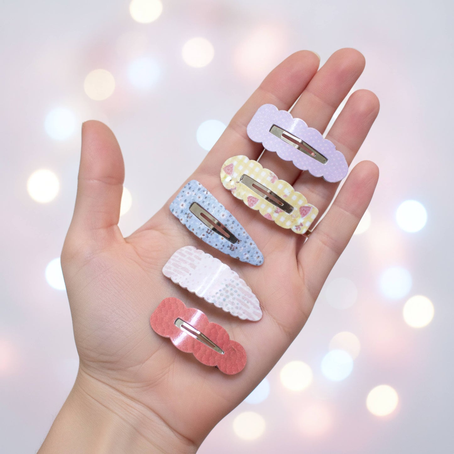 Five pastel hair claw clips in different colors and patterns are displayed on an open hand against a soft, dreamy bokeh background of warm lights.