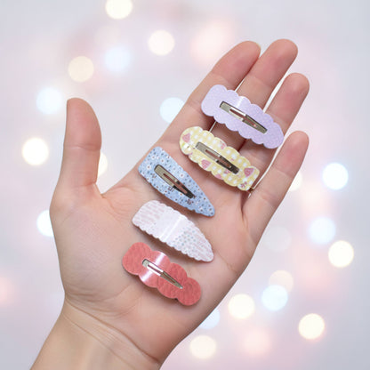 Five pastel hair claw clips in different colors and patterns are displayed on an open hand against a soft, dreamy bokeh background of warm lights.