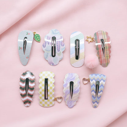 Eight decorative hair clips arranged on soft pink fabric. The clips feature pastel patterns including plaid, chevron, floral, and iridescent finishes, with small charms such as a leaf, star, heart, and pom-pom.