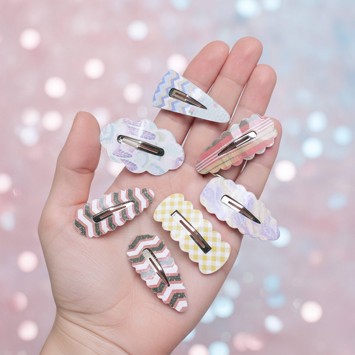 Six pastel patterned hair claw clips held in a person's hand against a soft pink and blue bokeh background.