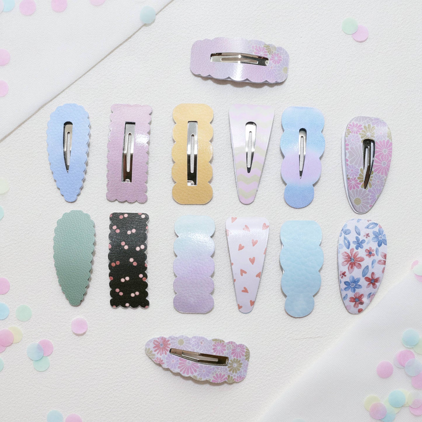 Twelve pastel hair claw clips arranged on a white textured background with scattered pastel confetti. The clips come in multiple colors and patterns including solid pastel blue, pink, yellow, mint green, black with pink dots, lavender, white with pink hearts, and floral prints.