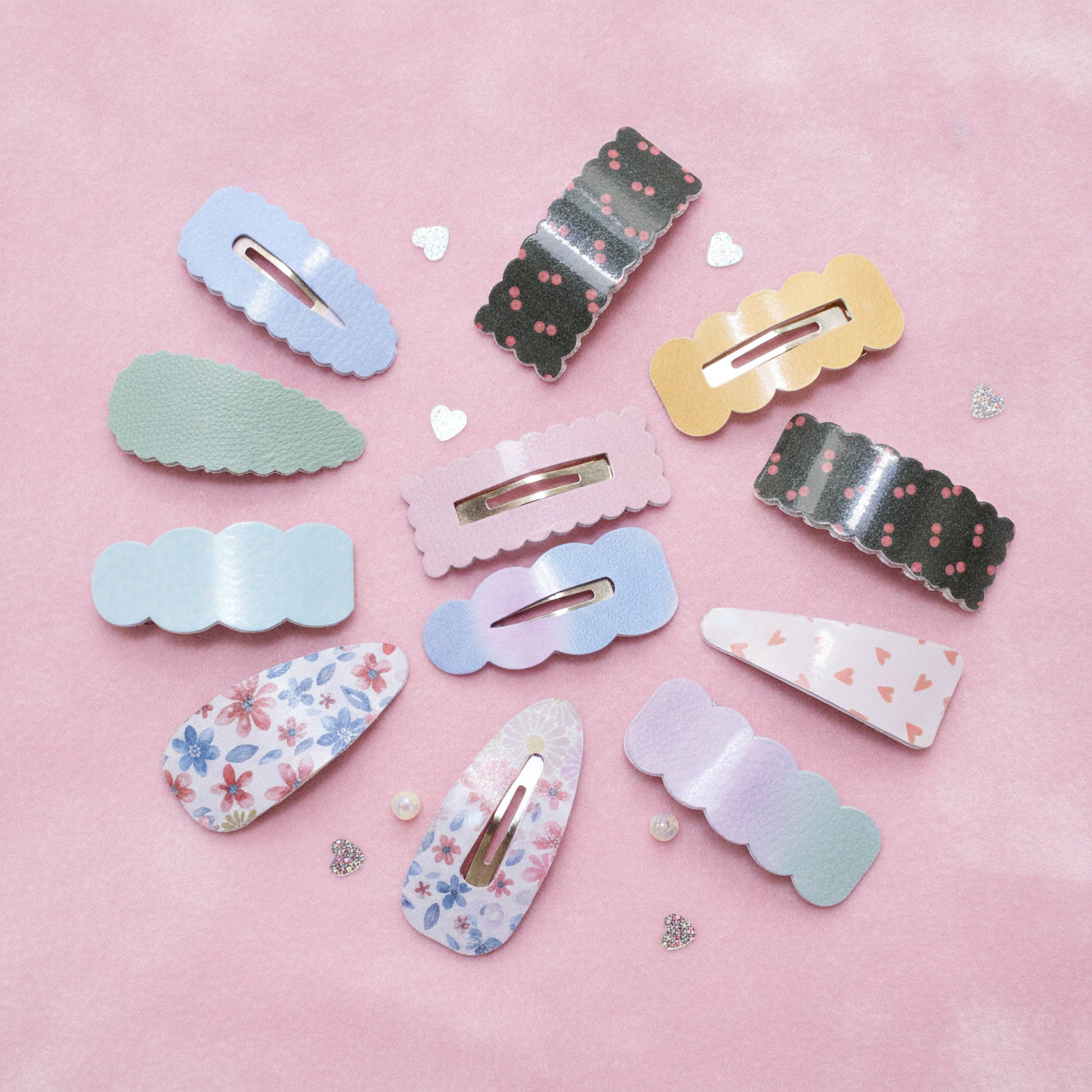 A set of twelve pastel-colored hair clips with various patterns and scalloped edges, arranged on a soft pink background with small decorative hearts and glitter.