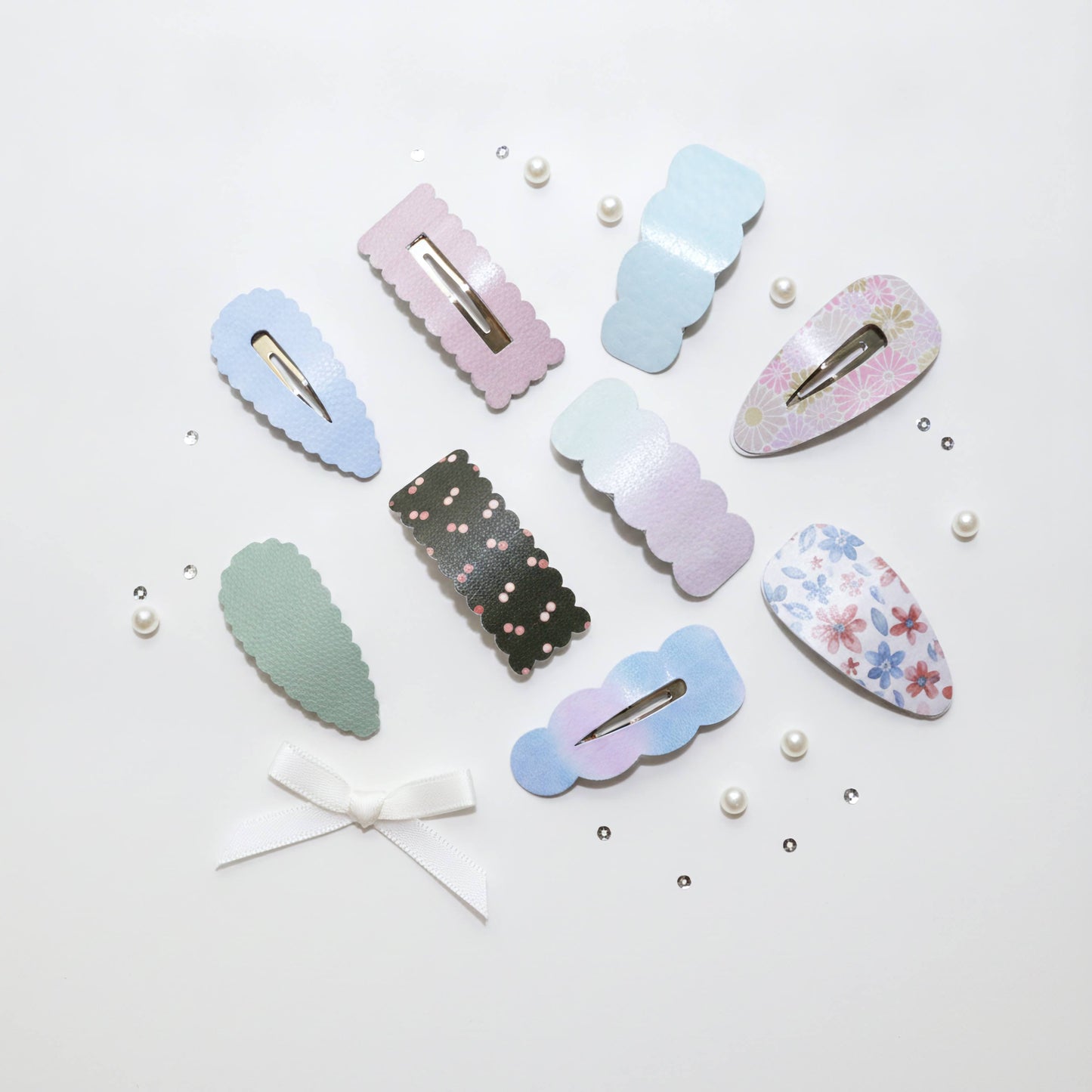 A set of pastel hair claw clips arranged on a white background with decorative pearl beads and small rhinestones. The clips come in various shapes and patterns, including solid pastel blue, pink, mint green, black with pink dots, iridescent lavender, floral print, and a white ribbon bow clip.
