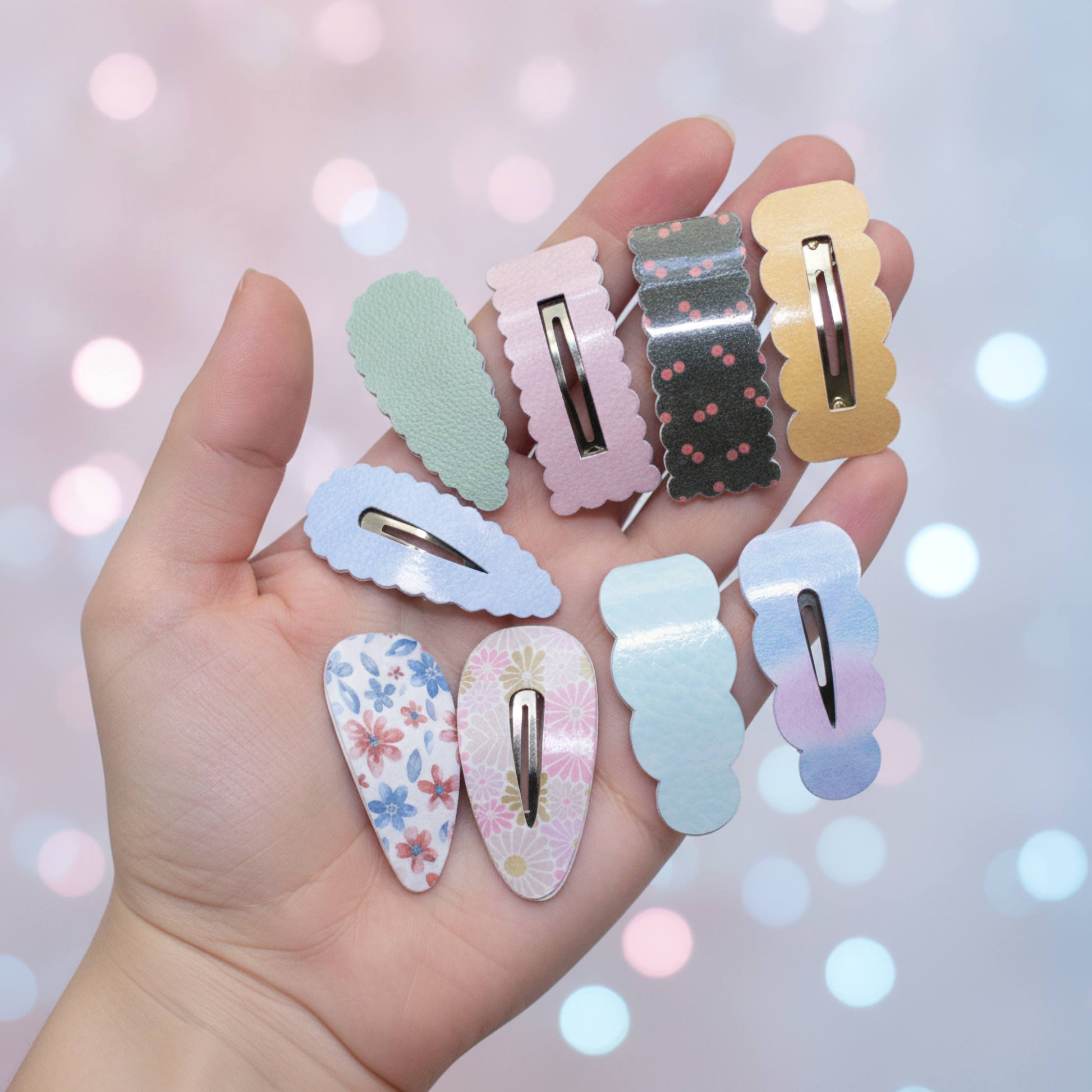 A hand holds eight decorative hair claw clips in pastel colors and patterns against a soft pink bokeh background. The clips include solid pastel shades, floral prints, and dotted patterns.