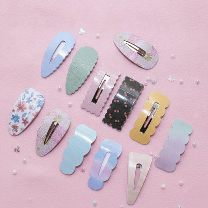 A set of pastel hair claw clips arranged on a pink background with small decorative pearls and glitter. The clips feature various patterns including floral prints, polka dots, ombre gradients, and solid colors in blue, green, pink, black, gold, and purple.