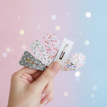 A hand holds five decorative hair clips against a pastel pink-to-blue background with soft bokeh lights. The clips have scalloped edges and different patterns: gray with white polka dots, white with small pink hearts, white with small blue hearts, white with small multicolor dots, and white with small pink and blue dots.
