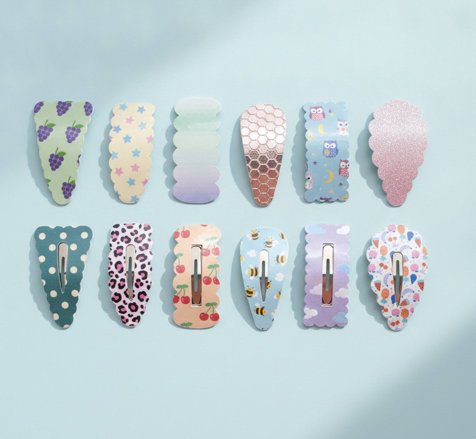 Ten decorative hair claw clips arranged in two rows on a pale blue background. The clips feature various pastel patterns including grapes, stars, gradient, honeycomb, owls, glitter, polka dots, leopard print, cherries, bees, clouds, and floral motifs.