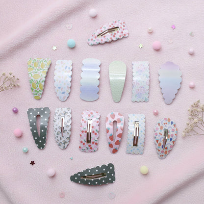 Twelve pastel hair claw clips arranged in rows on a soft pink fabric background with small decorative beads and dried flowers. The clips feature various pastel patterns including floral, polka dots, hearts, and gradients in shades of pink, blue, green, and white.