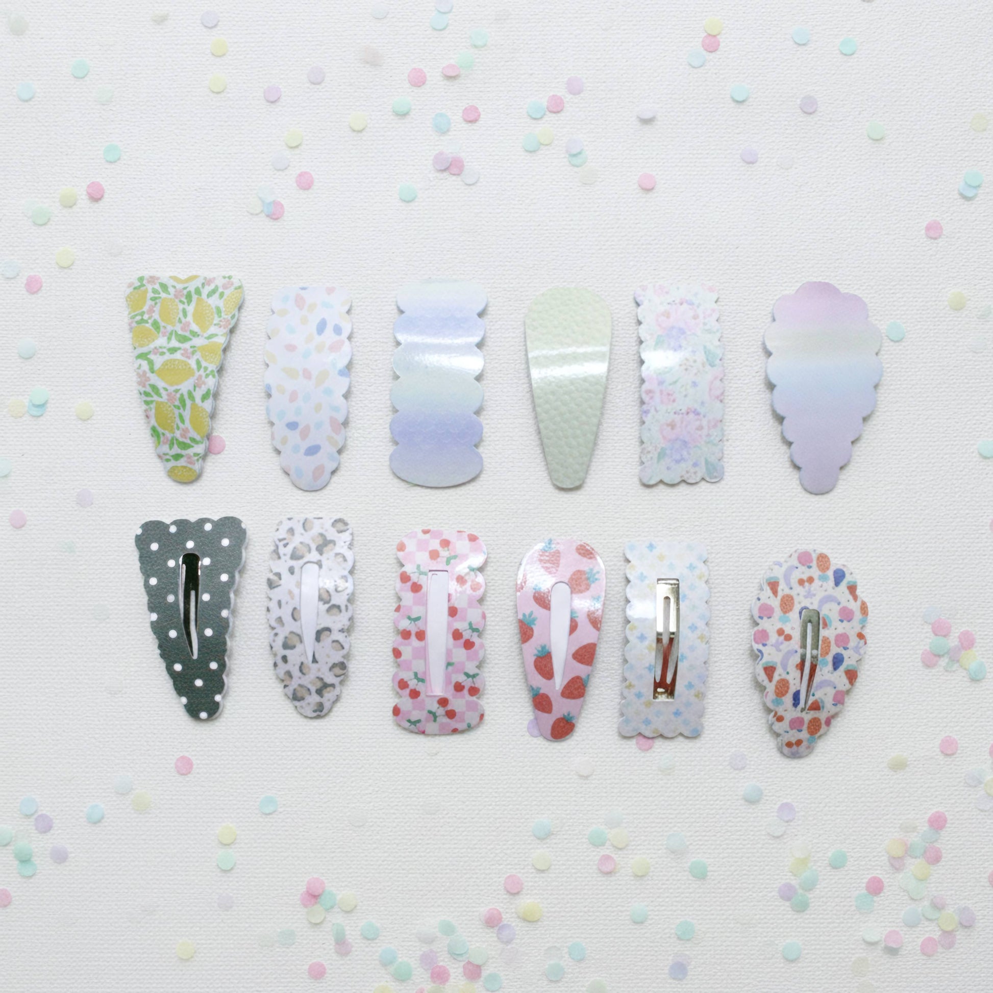 Twelve decorative hair claw clips arranged in two rows on a white textured background with scattered pastel confetti. The clips feature various pastel patterns including lemonade, polka dots, watercolor gradients, and fruit prints.