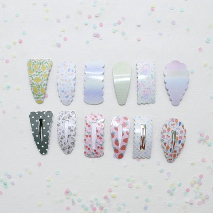 Twelve decorative hair claw clips arranged in two rows on a white textured background with scattered pastel confetti. The clips feature various pastel patterns including lemonade, polka dots, watercolor gradients, and fruit prints.