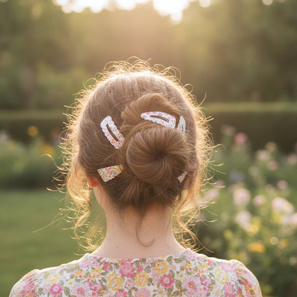A woman viewed from behind in a sunlit garden, wearing three white floral hair clips in her hair bun.