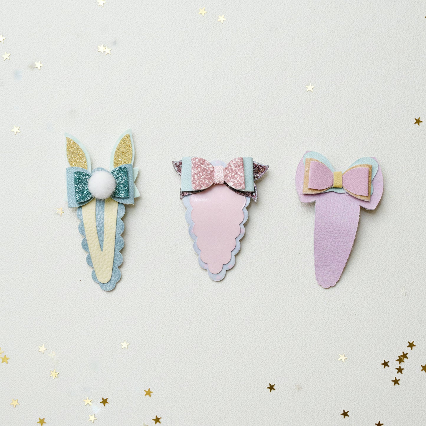Three pastel hair claw clips arranged on a light cream background with scattered gold star confetti. The left clip is cream and teal with glittery gold ears and a white pom-pom.