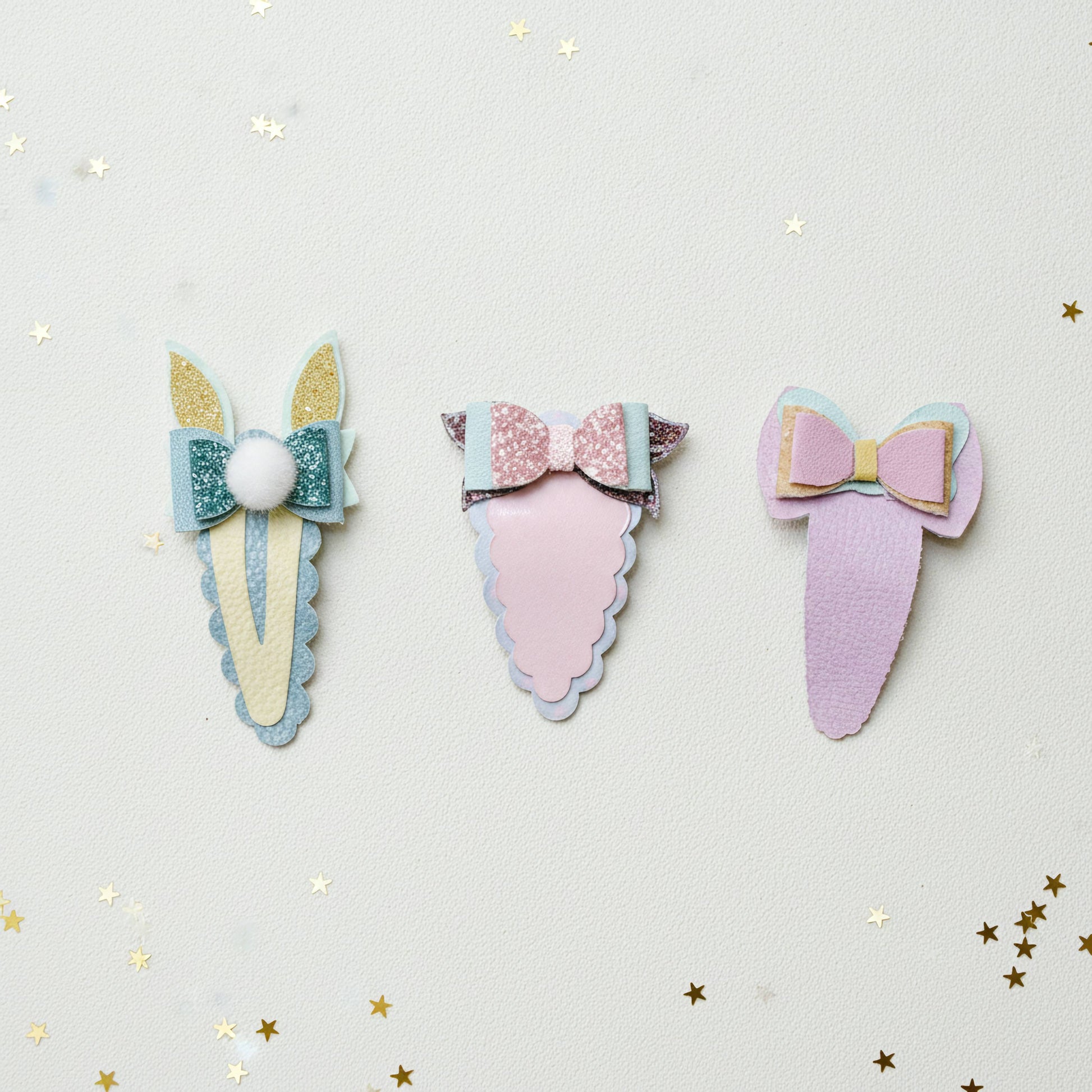 Three pastel hair claw clips arranged on a light cream background with scattered gold star confetti. The left clip is cream and teal with glittery gold ears and a white pom-pom.