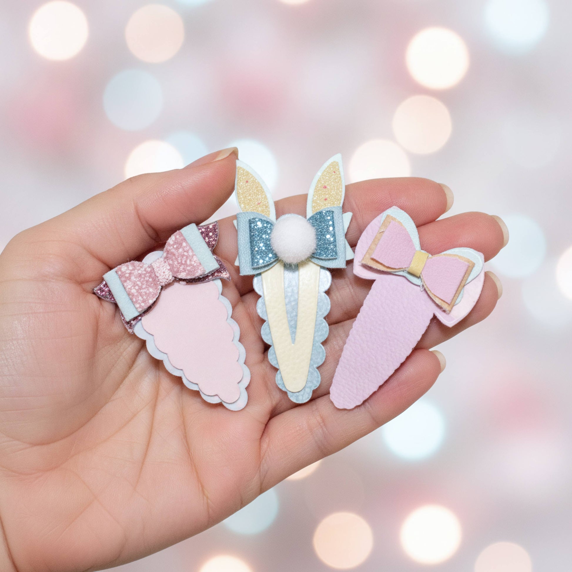 Three pastel bunny-themed hair clips held in a hand against a soft pink and white bokeh background. The clips feature light pink bunny bodies with glittery bows in pink, blue, and gold, and a central white pom-pom.