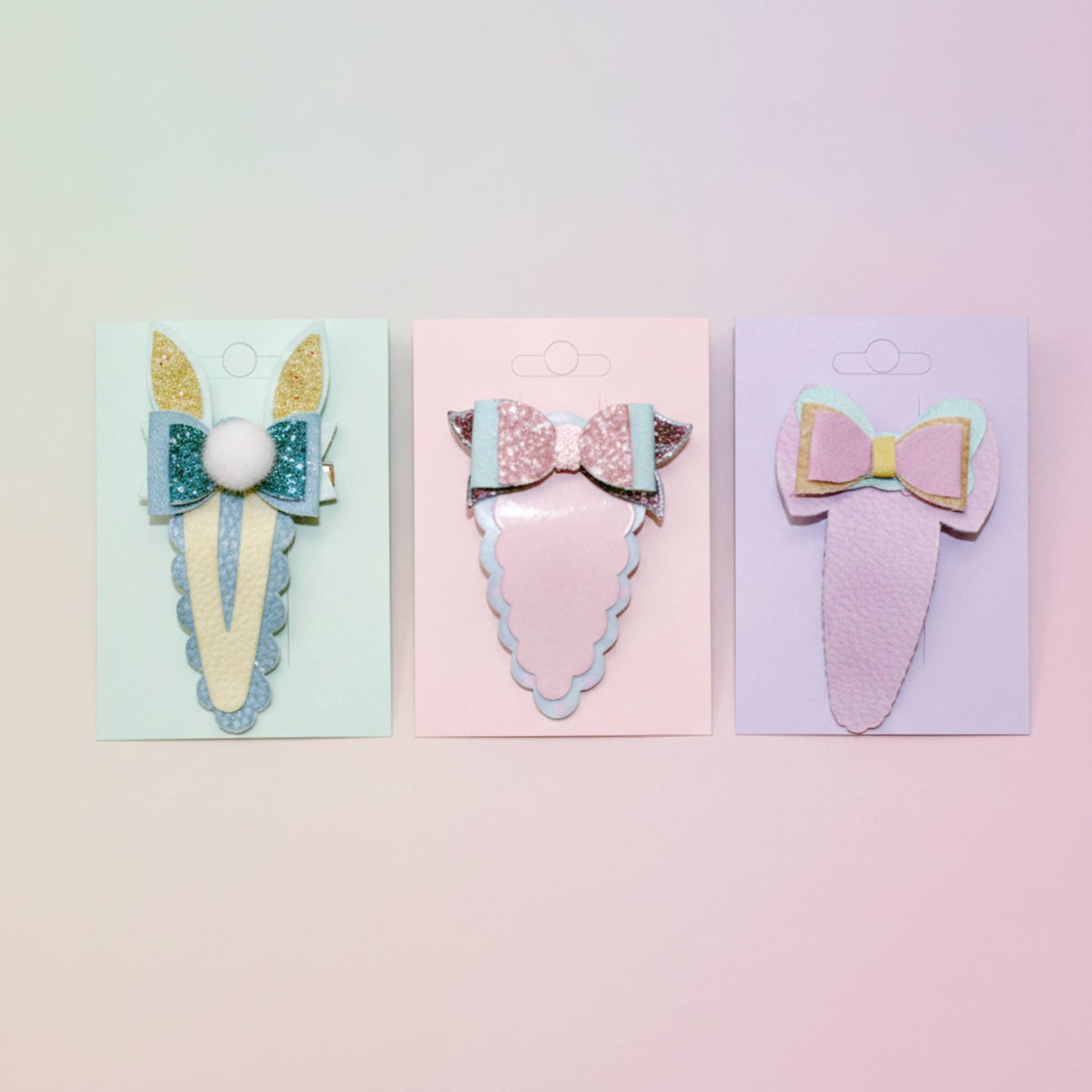 Three pastel-colored hair clips on individual display cards against a soft pink and yellow gradient background. The clips feature whimsical designs including bunny ears, a glittery bow with a pom-pom, and a bow with layered pastel wings.