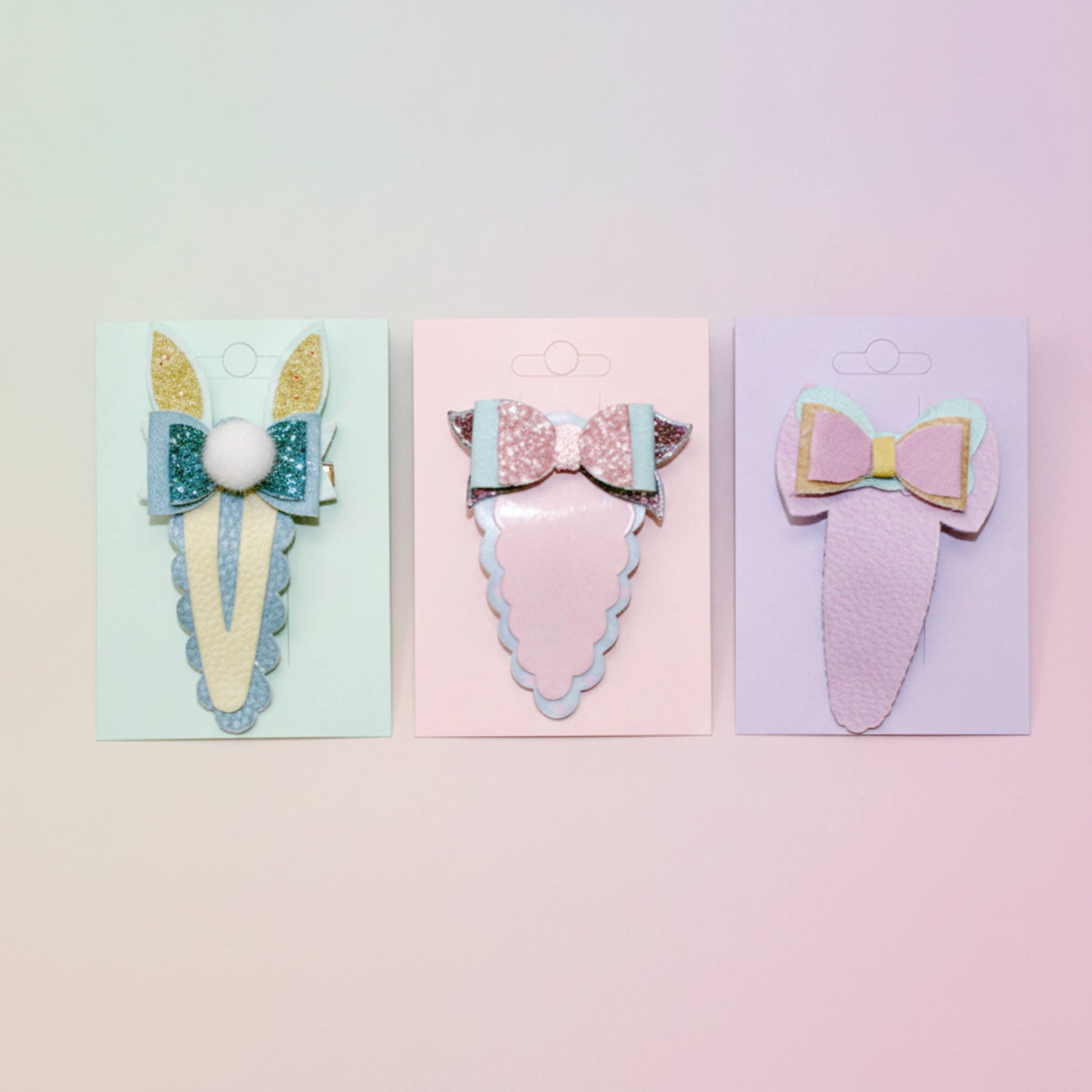 Three pastel-colored hair clips on individual display cards against a soft pink and yellow gradient background. The clips feature whimsical designs including bunny ears, a glittery bow with a pom-pom, and a bow with layered pastel wings.