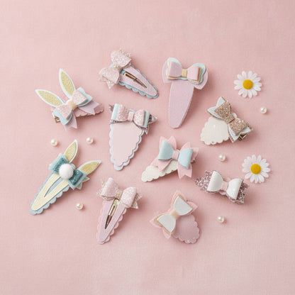 A set of pastel hair clips arranged on a soft pink background, including bow, bunny-ear, scalloped, and glitter styles in pink, blue, white, and gold tones, with small daisy props and pearl beads around them.