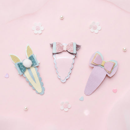 Three pastel hair clips arranged on a soft pink background with small decorative flowers and pearl-like beads. The clips have bow and bunny-ear designs in pastel colors including pink, lavender, blue, and yellow.