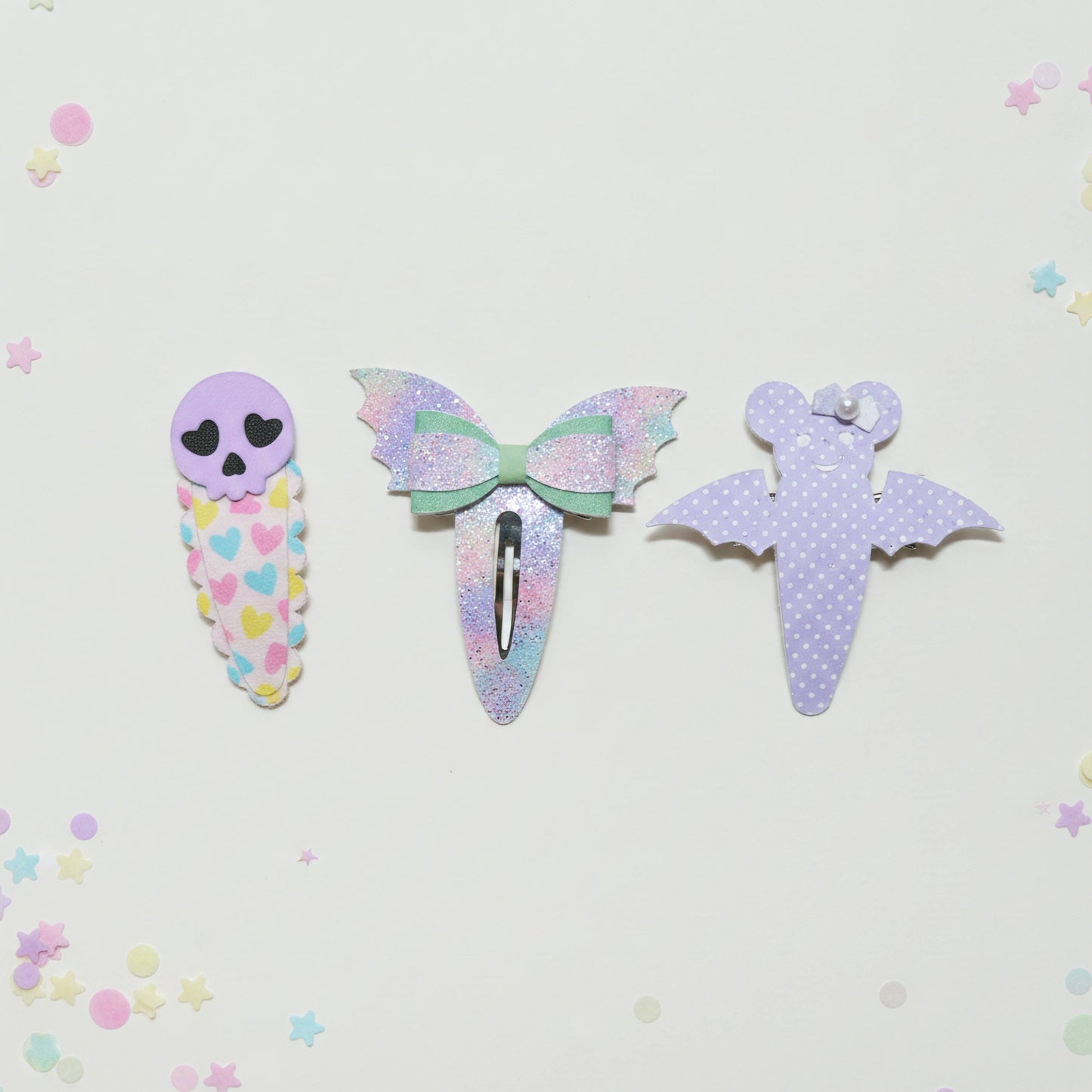 Three cute pastel hair clips arranged on a white background with scattered star confetti. The left clip is lavender with a skull face and multicolor heart pattern.
