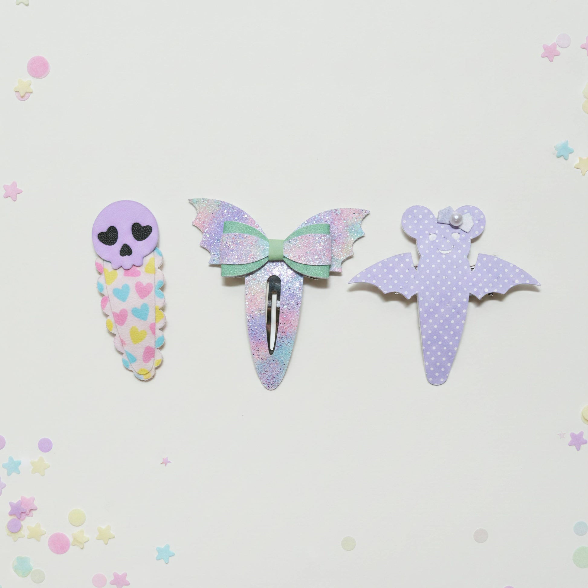 Three cute pastel hair clips arranged on a white background with scattered star confetti. The left clip is lavender with a skull face and multicolor heart pattern.