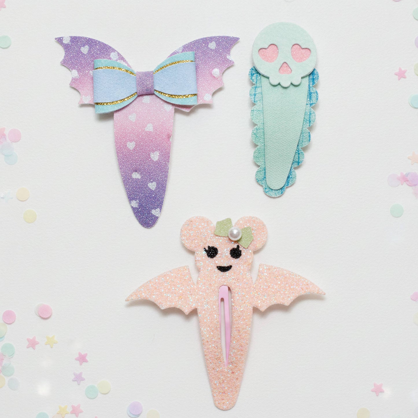 Three cute pastel hair clips styled like a bow, a skull, and a bat, arranged on a white background with colorful confetti.