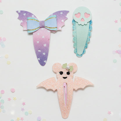 Three cute pastel hair clips styled like a bow, a skull, and a bat, arranged on a white background with colorful confetti.