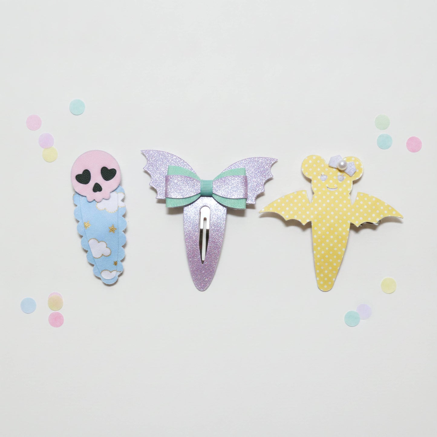 Three cute hair clips styled like a skeleton baby, a glittery mermaid bow, and a yellow polka-dot bat, arranged on a white background with pastel confetti.