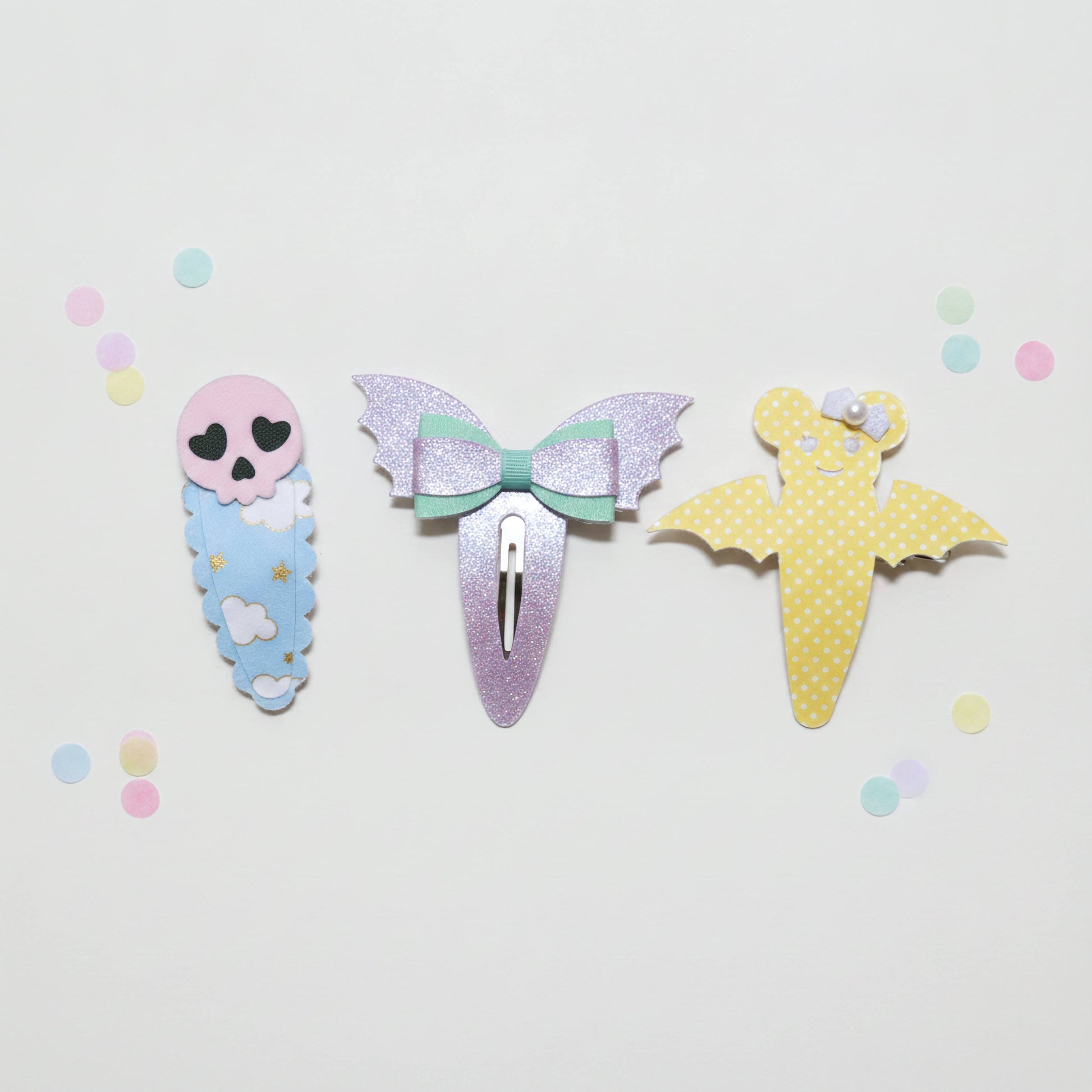 Three cute hair clips styled like a skeleton baby, a glittery mermaid bow, and a yellow polka-dot bat, arranged on a white background with pastel confetti.