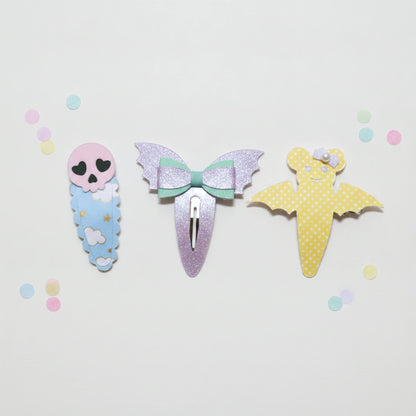 Three cute hair clips styled like a skeleton baby, a glittery mermaid bow, and a yellow polka-dot bat, arranged on a white background with pastel confetti.