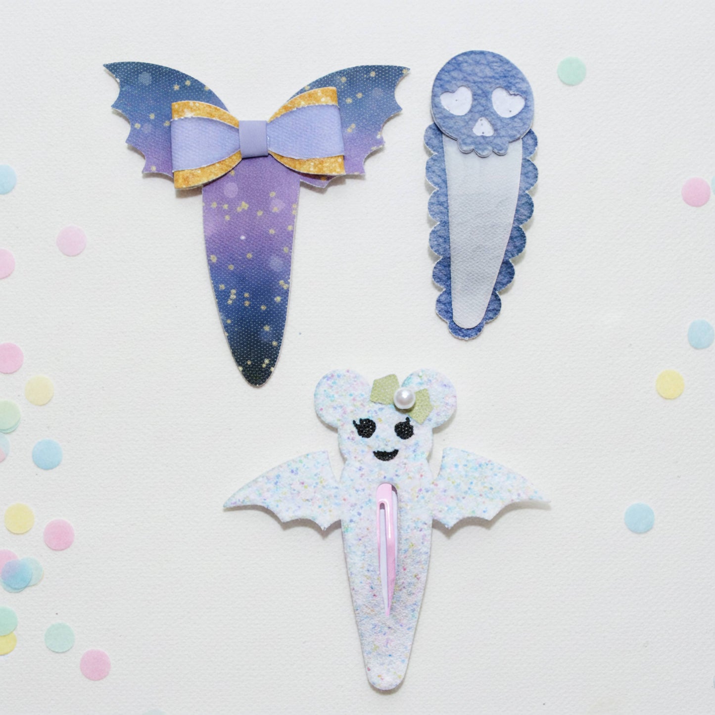 Three cute ghost-themed hair clips arranged on a white background with pastel confetti. The top-left clip is purple and blue with gold glitter stars and a lavender bow.