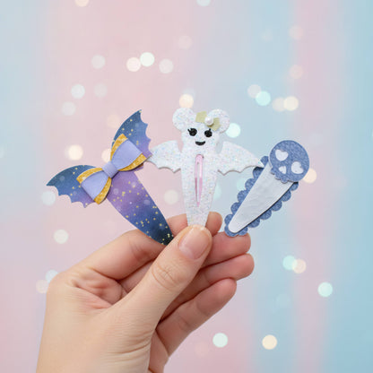 A hand holds three cute ghost-themed hair clips against a pastel pink and blue bokeh background. The clips feature a purple bat-wing design with a lavender bow, a white glittery ghost, and a blue paw-print ice cream cone.